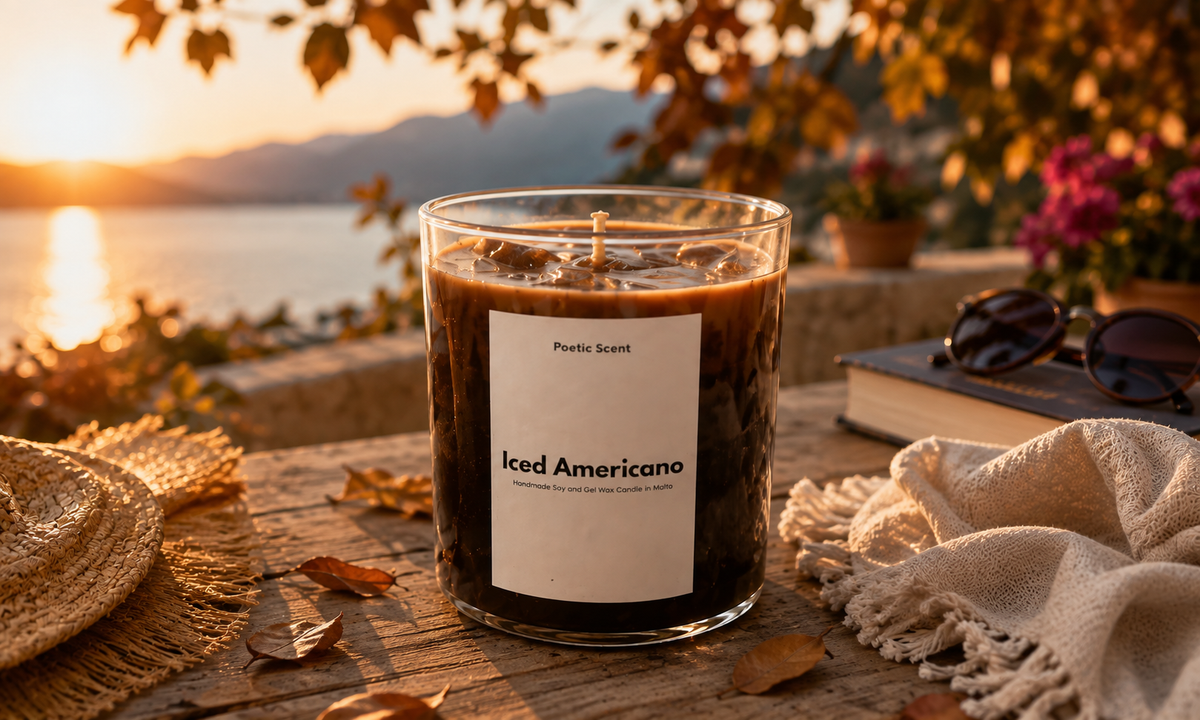 A glass jar candle labeled 'Iced Americano' with a white label and dark brown wax, styled on a wooden table outdoors at sunset with a lake, mountains, sunglasses, a straw hat, and a cream throw blanket in the background.